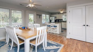 A dining area with a wooden table and white chairs, a kitchen with white cabinets, and a ceiling fan. - Sweet M'Ocean | Charming Edisto Beach Retreat: Steps to Shore,Covered Porch,Golf Cart Paradise (Edisto Island)