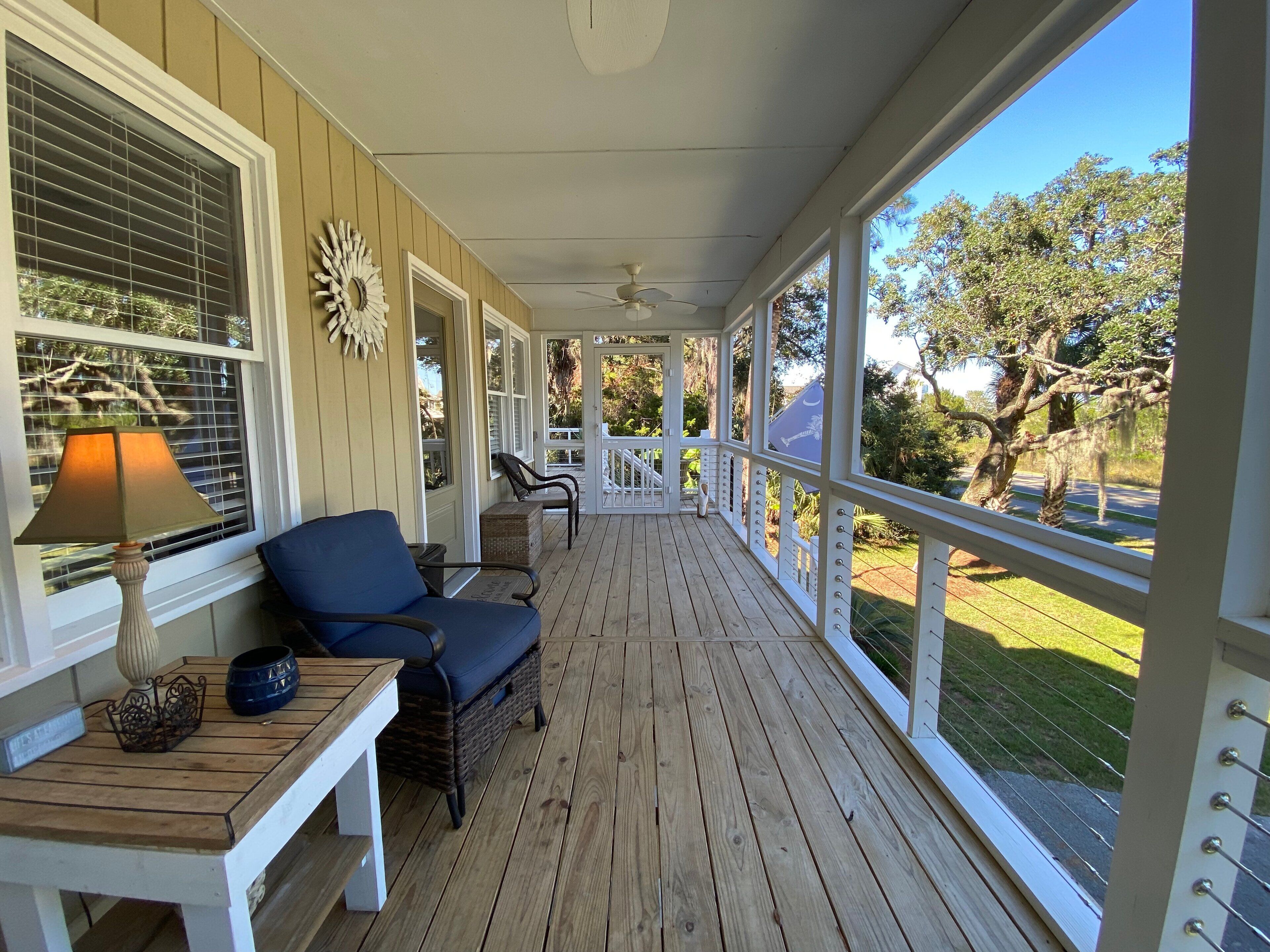 A covered porch with a wooden floor, a blue chair, a small table with a lamp, and a decorative wall piece.