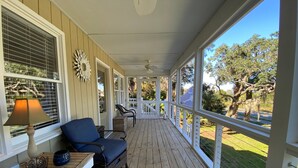 A covered porch with a wooden floor, a blue chair, a small table with a lamp, and a decorative wall piece. - Sweet M'Ocean | Charming Edisto Beach Retreat: Steps to Shore,Covered Porch,Golf Cart Paradise (Edisto Island)