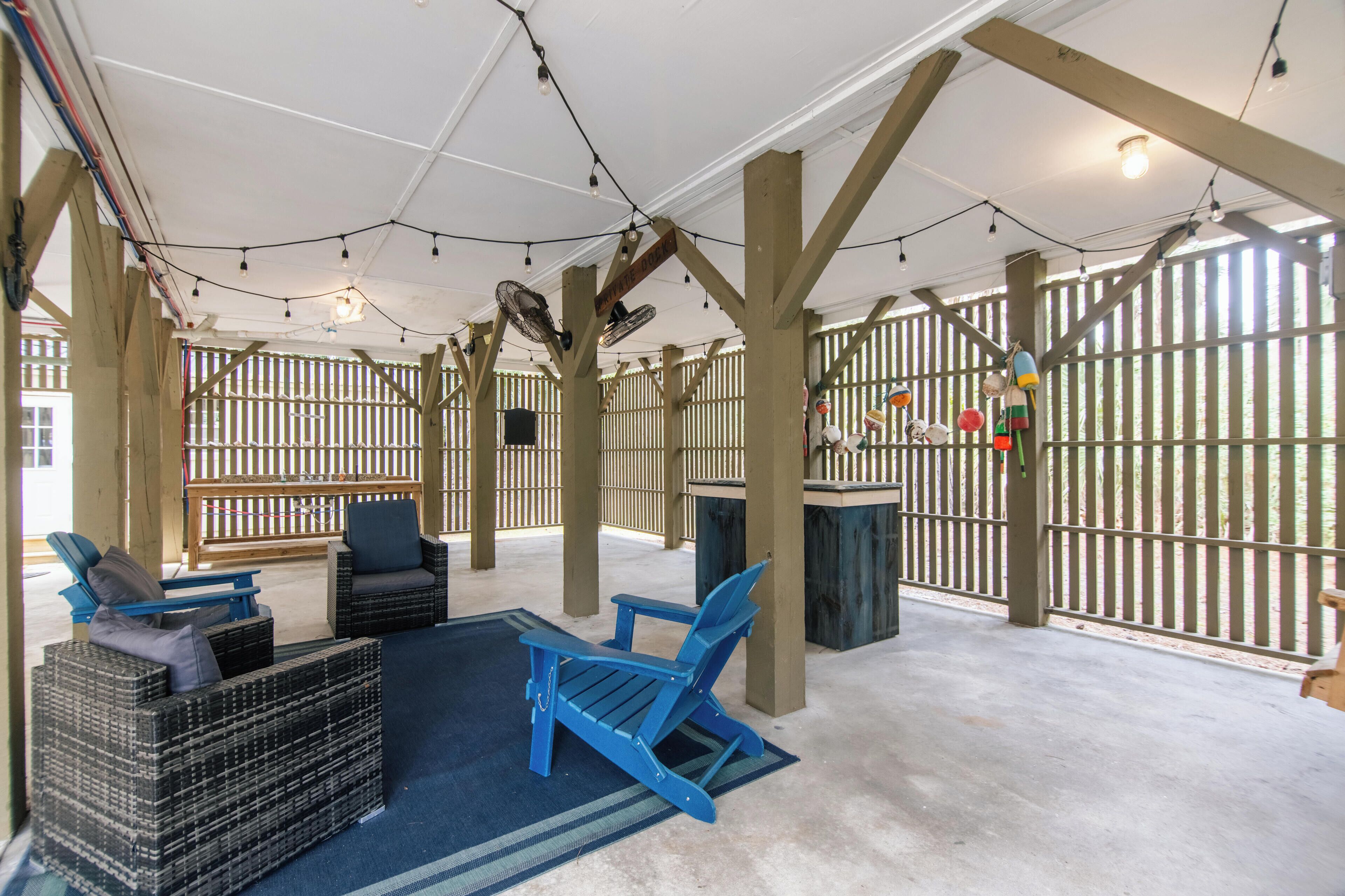 An outdoor seating area with blue chairs and a wicker sofa, wooden beams, string lights, and a view through a wooden fence.