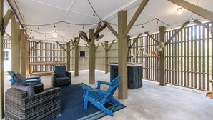 An outdoor seating area with blue chairs and a wicker sofa, wooden beams, string lights, and a view through a wooden fence. - Sweet M'Ocean | Charming Edisto Beach Retreat: Steps to Shore,Covered Porch,Golf Cart Paradise (Edisto Island)