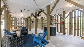 An outdoor seating area with blue chairs and a wicker sofa, wooden beams, string lights, and a view through a wooden fence.