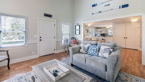 A spacious living room with a grey sofa, a coffee table, and a dining area with a kitchen in the background. - Sweet M'Ocean | Charming Edisto Beach Retreat: Steps to Shore,Covered Porch,Golf Cart Paradise (Edisto Island)