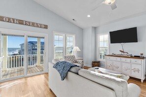 A spacious living room with a large window, a flat-screen TV, and a sign reading "OAK ISLAND". - Crasea: Ocean Views and Fenced Backyard Oasis with Pool! (Oak Island)