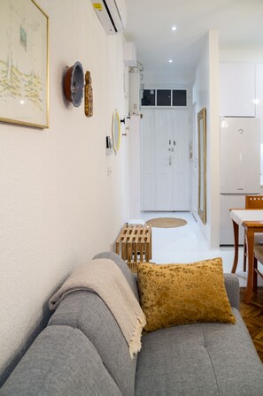 Uma sala de estar moderna com um sofá cinza, uma almofada decorativa estampada e uma mesa de centro de madeira. Uma porta branca dá acesso a uma cozinha com uma geladeira branca e uma mesa de jantar.