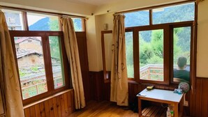 A room with wooden floors, a bed with a red blanket, a small table with a fan, and large windows with curtains. - Private Room for long and short stay (Manali)