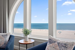 A room with a large window offering a beach view, a small round table with a plant, and blue upholstered furniture. - 🏖️ Gorgeous Ocean Front Belmont Towers Just Renovated, Pool, Gym. on Boardwalk 🏖️ (Ocean City)
