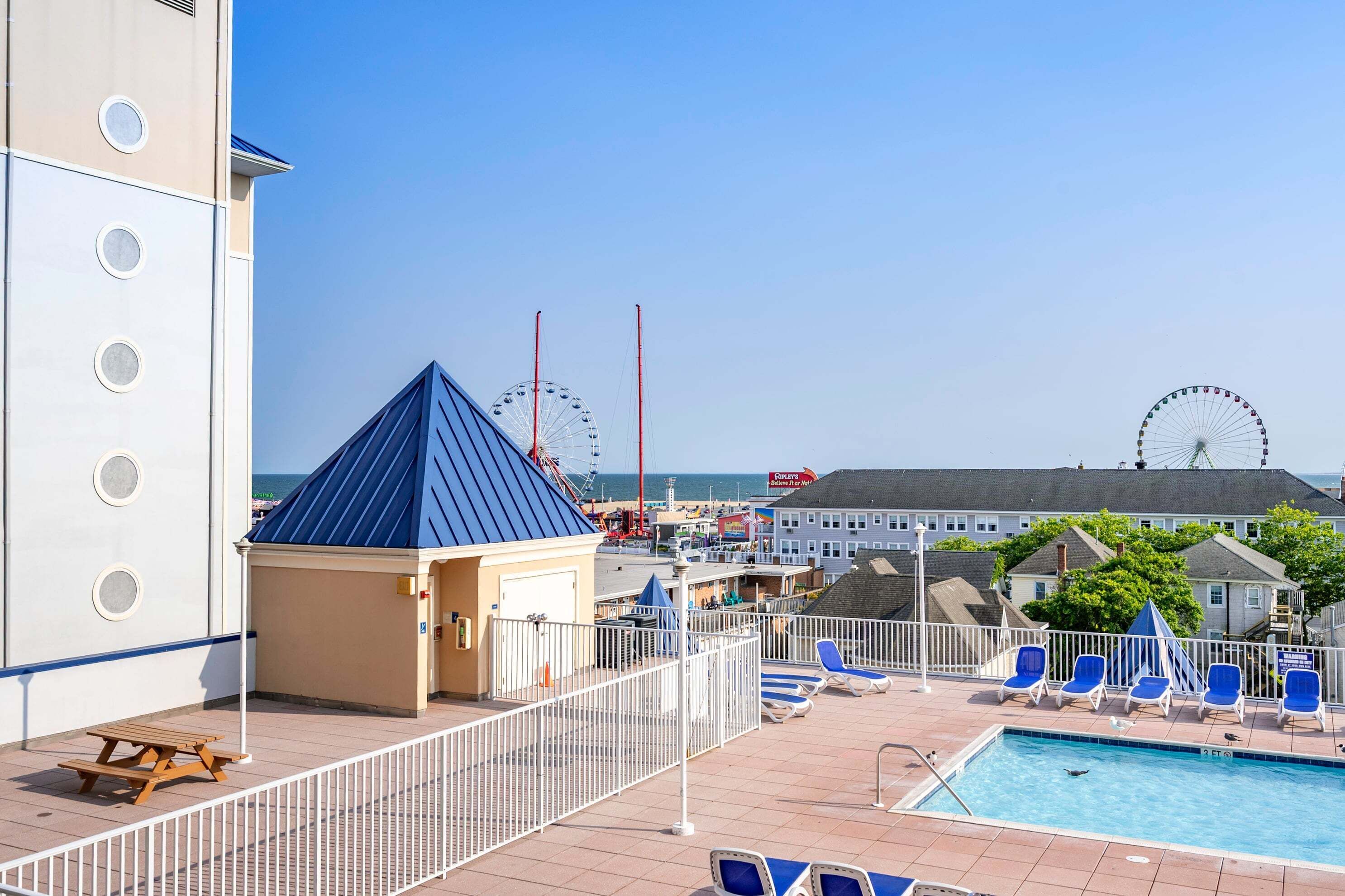 A rooftop pool area with lounge chairs, a small pavilion, and a view of the beach and ferris wheel.