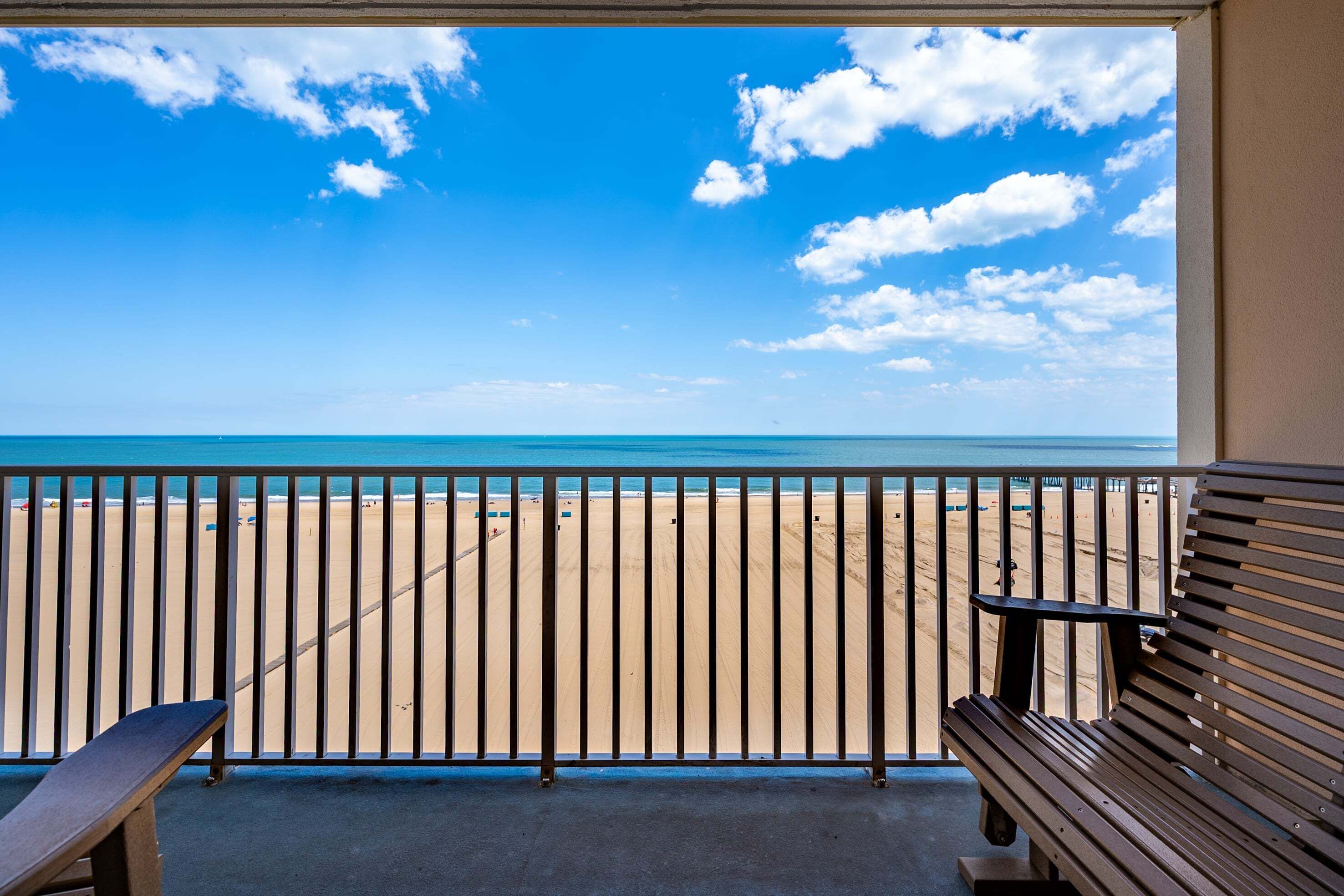 A balcony with a view of a beach and ocean, a bench, and a table.