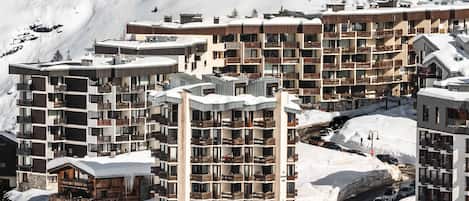 A snow-covered mountain resort with multi-story buildings, including a hotel with balconies and a ski lift visible in the background.