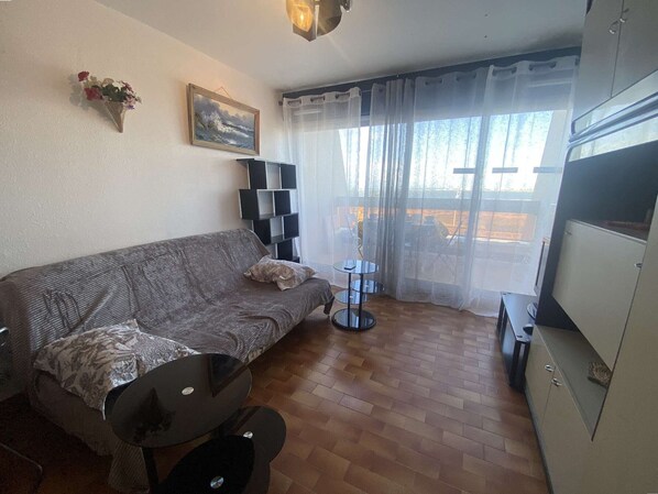 A living room with a sofa, a small round table, a bookshelf, and a window with sheer curtains. - Cozy studio for 4, beach access, free parking Port Camargue (Port Camargue)