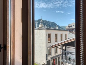 Vista a través de una ventana de un edificio con balcón, un cartel que dice 'GALERIE SAINT' y una montaña con una iglesia al fondo.