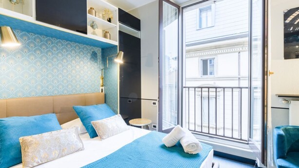 Ein Hotelzimmer mit Bett, einem kleinen Tisch, einem Sessel, einem Balkon mit Geländer und einem Fenster.