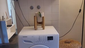 Een wasmachine met voorlaadklep, een bedieningspaneel, een klein houten krukje en een rieten mand ernaast.