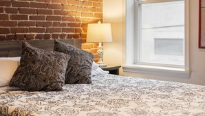 Een net opgemaakt bed met beddengoed met een patroon en twee decoratieve kussens, een bedlampje en een bakstenen muur.