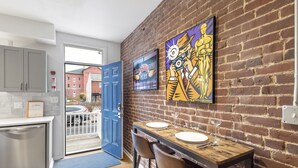 Een moderne keuken met een blauwe deur die leidt naar een balkon, een eethoek met twee stoelen en een tafel voor twee personen, en een metselwerk muur met een kunstwerk in een lijst.