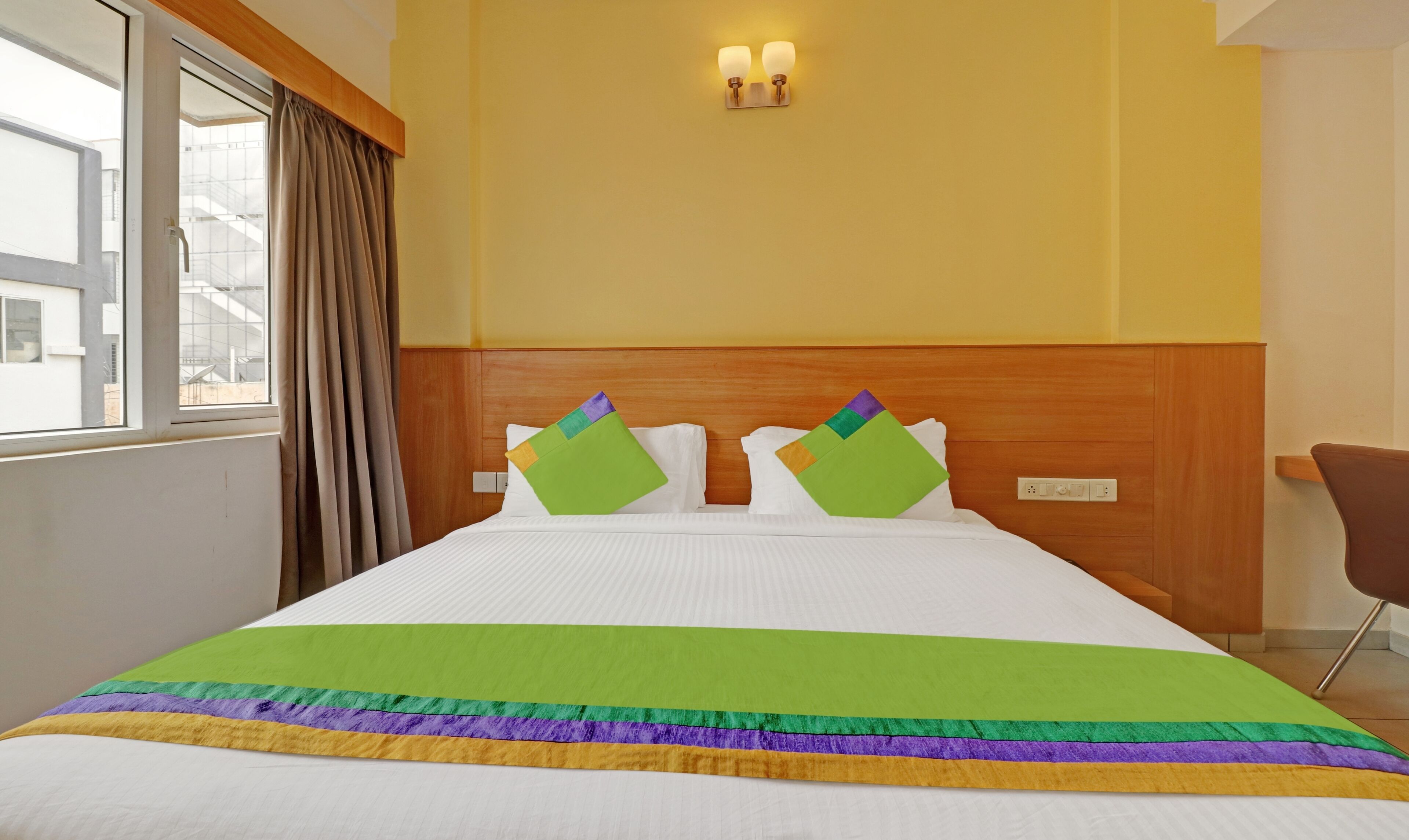 Quarto de hotel com uma cama grande, cabeceira de madeira, janela com cortinas e luminária fixada na parede.