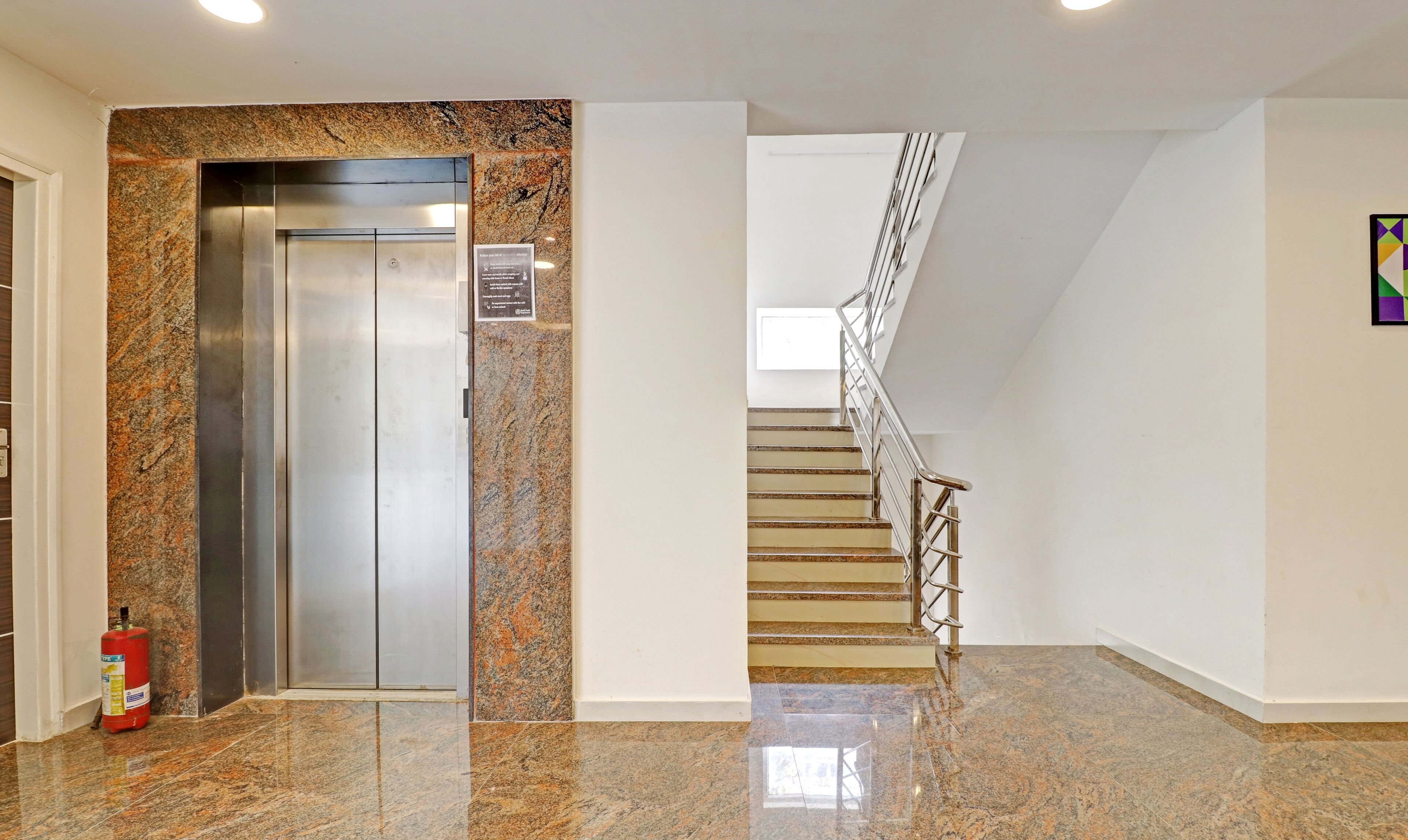 Un ascensor con pared de granito, un extintor y una escalera con barandillas de metal.