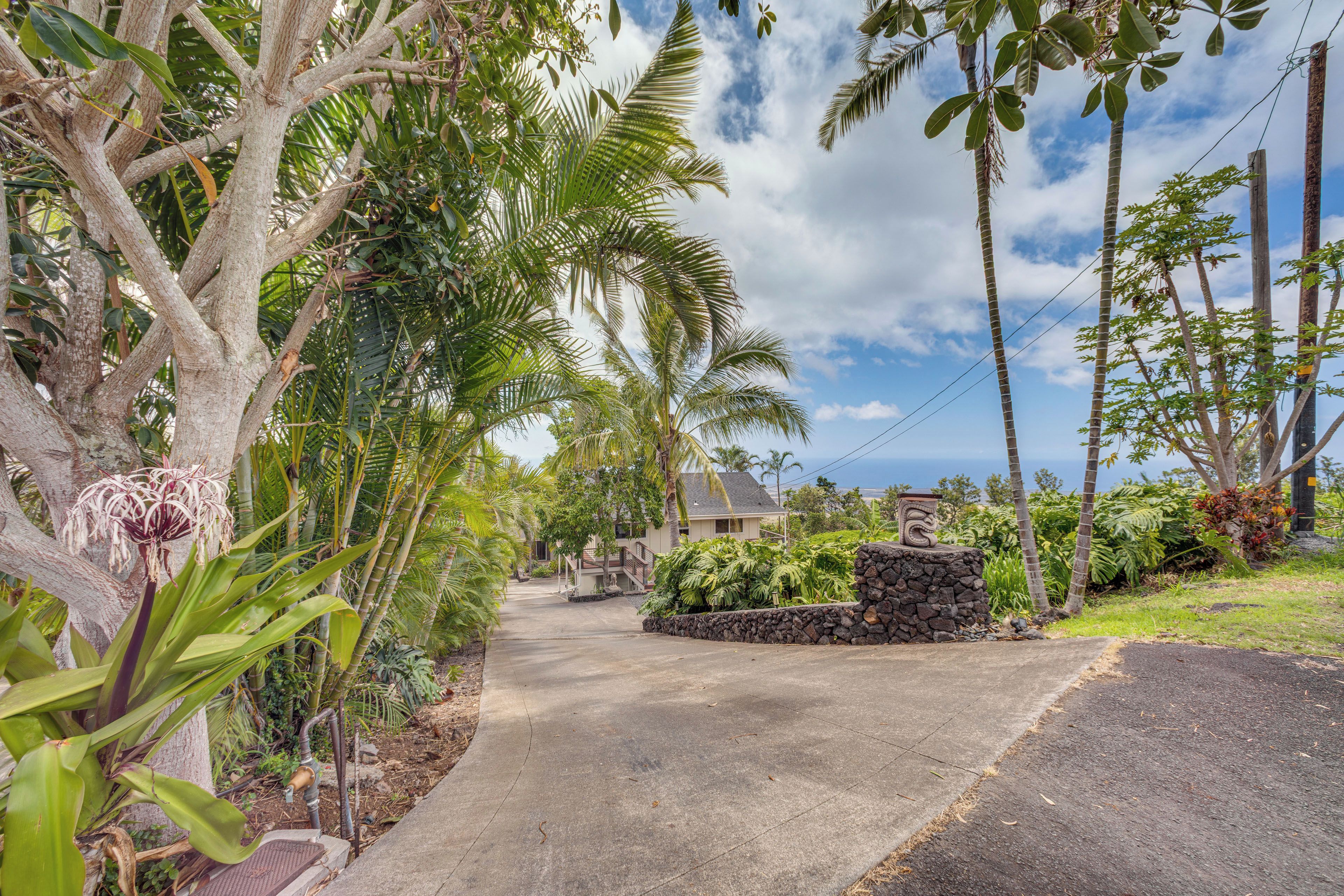 Un barrio residencial con entrada pavimentada, un muro de piedra y diversa vegetación tropical.