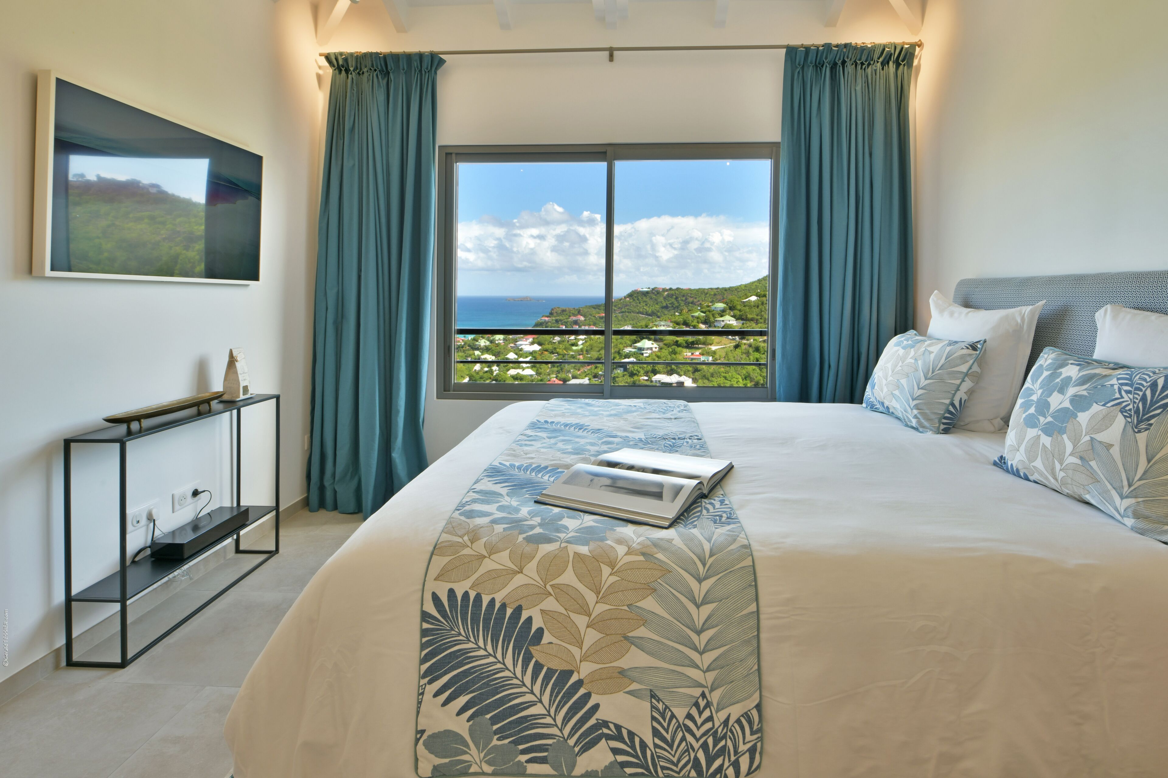 A bedroom with a large bed, a view of the sea, and a console table with a laptop.