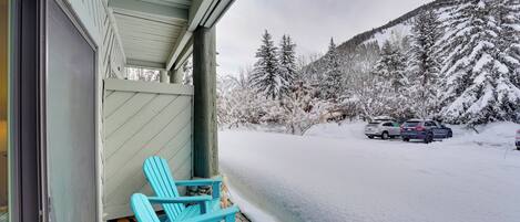 Varanda coberta com cadeiras Adirondack azuis, um caminho de acesso coberto de neve e montanhas ao fundo.