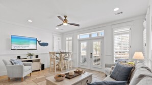 A spacious living room with a large TV, comfortable seating, a ceiling fan, and large windows.