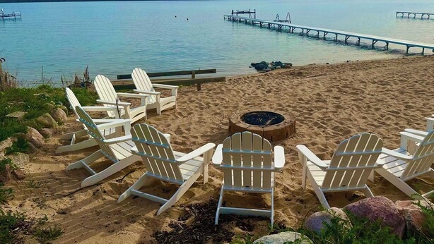 Een strand met witte Adirondack-stoelen, een vuurkorf en een houten steiger die zich uitstrekt over kalm water.