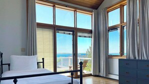 Een slaapkamer met een bed, een dressoir en grote ramen met uitzicht op zee.