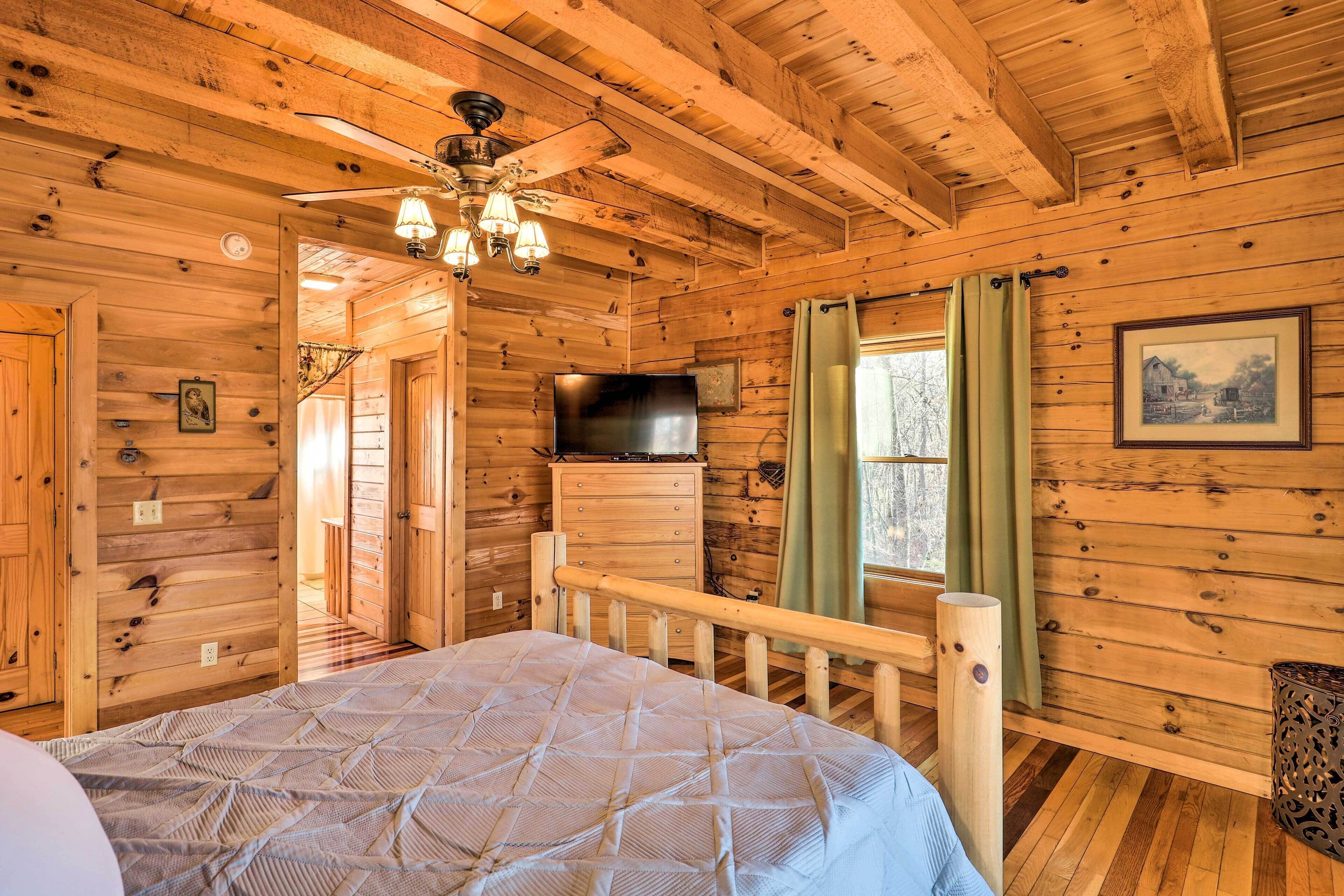 Interior de una cabaña de madera con cama, ventilador de techo, televisor montado y ventana con cortinas.