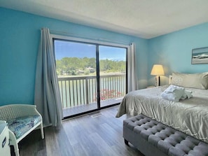 A bedroom with a large bed, a nightstand, a lamp, a bench, and a balcony with a view of greenery.