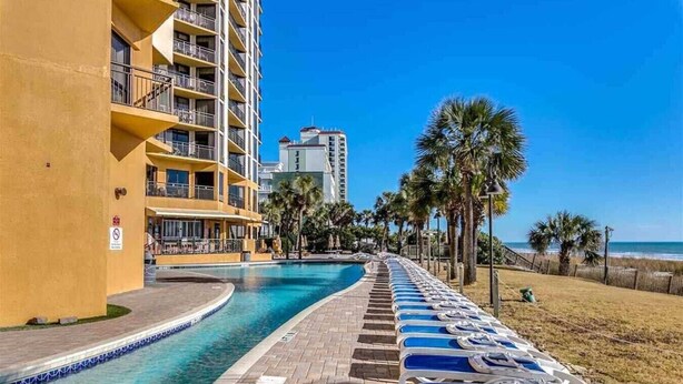 Un espace piscine d’hôtel offrant une vue dégagée sur l’océan, une promenade bordée de palmiers et un bâtiment doté de balcons.