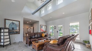 A spacious living room with a high ceiling, a fireplace, and a large window offering a view of the outdoors. - Glamping in Spicewood TX Hill Country~ Cabin 1 The Madison at Green Acres (Spicewood)