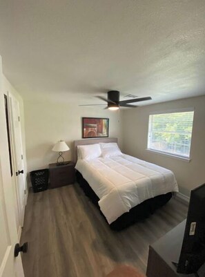 A bedroom with a bed, a ceiling fan, a window with blinds, a nightstand with a lamp, and a door.