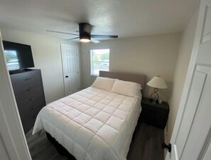 Un dormitorio con cama, tocador, televisor, ventilador de techo, ventana y mesita de noche con lámpara.