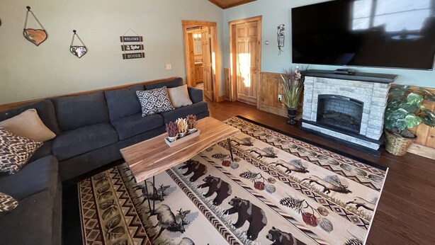 A cozy living room with a sectional sofa, a fireplace, a television, and a rug with animal prints. - Heart Lodge,slg fam sleeps 11, Lakefront great swimming / fishing kayaks, padbd. (Orland)