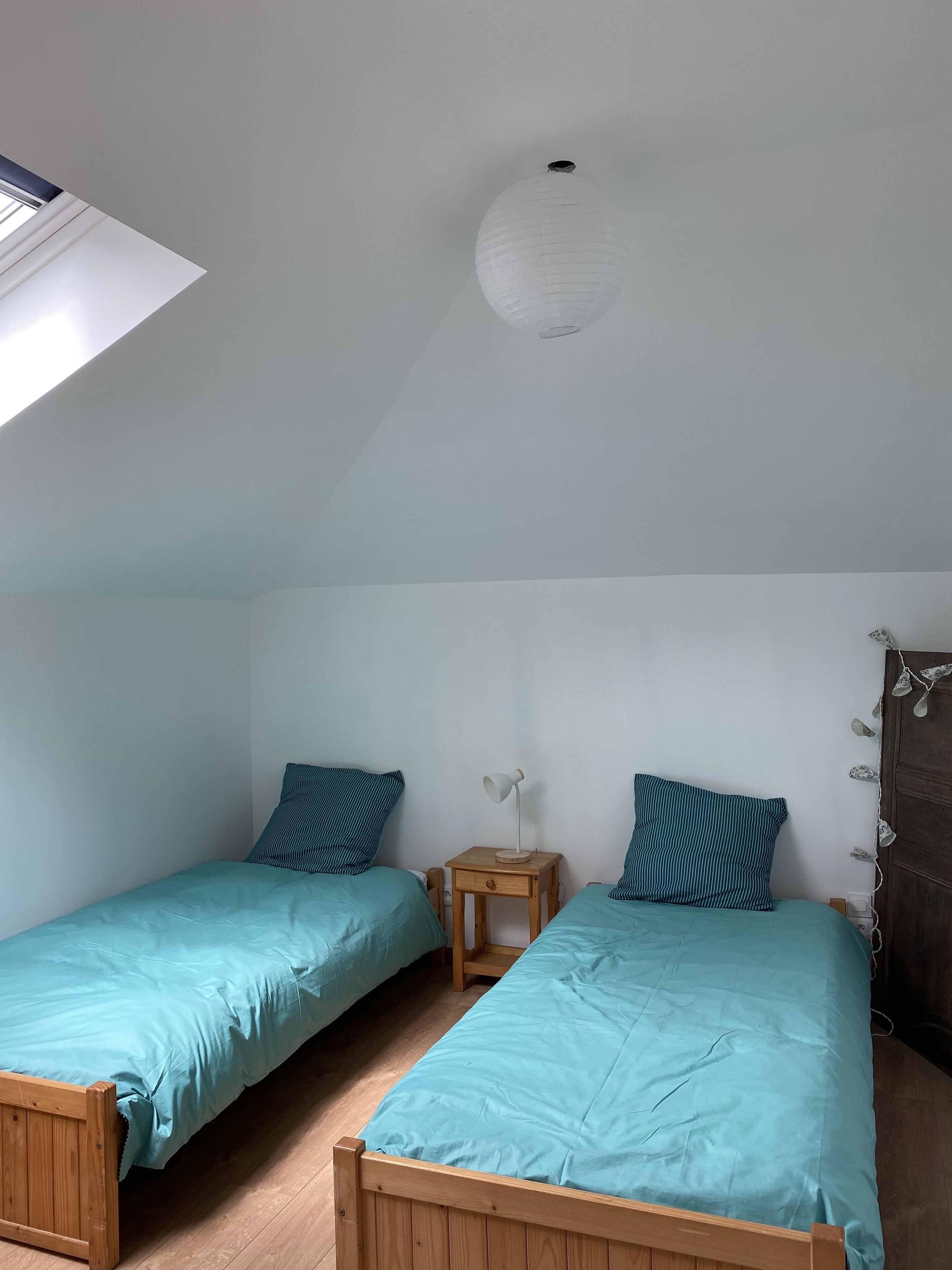 Una habitación con dos camas de madera, cada una con colcha color turquesa y almohadas a rayas, una mesita de noche de madera con lámpara, y un candelabro en el techo blanco.