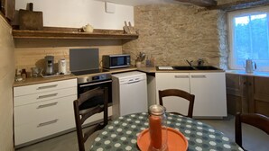 Une cuisine de taille réduite avec un mur en pierre, des poutres en bois, une table à manger recouverte d’une nappe à motifs et une seule chaise.