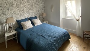 Une chambre avec une couvre-lit bleue, des oreillers blancs et bleus, deux tables de chevet avec des lampes, une fenêtre avec des rideaux légers et un papier peint à motifs floraux.
