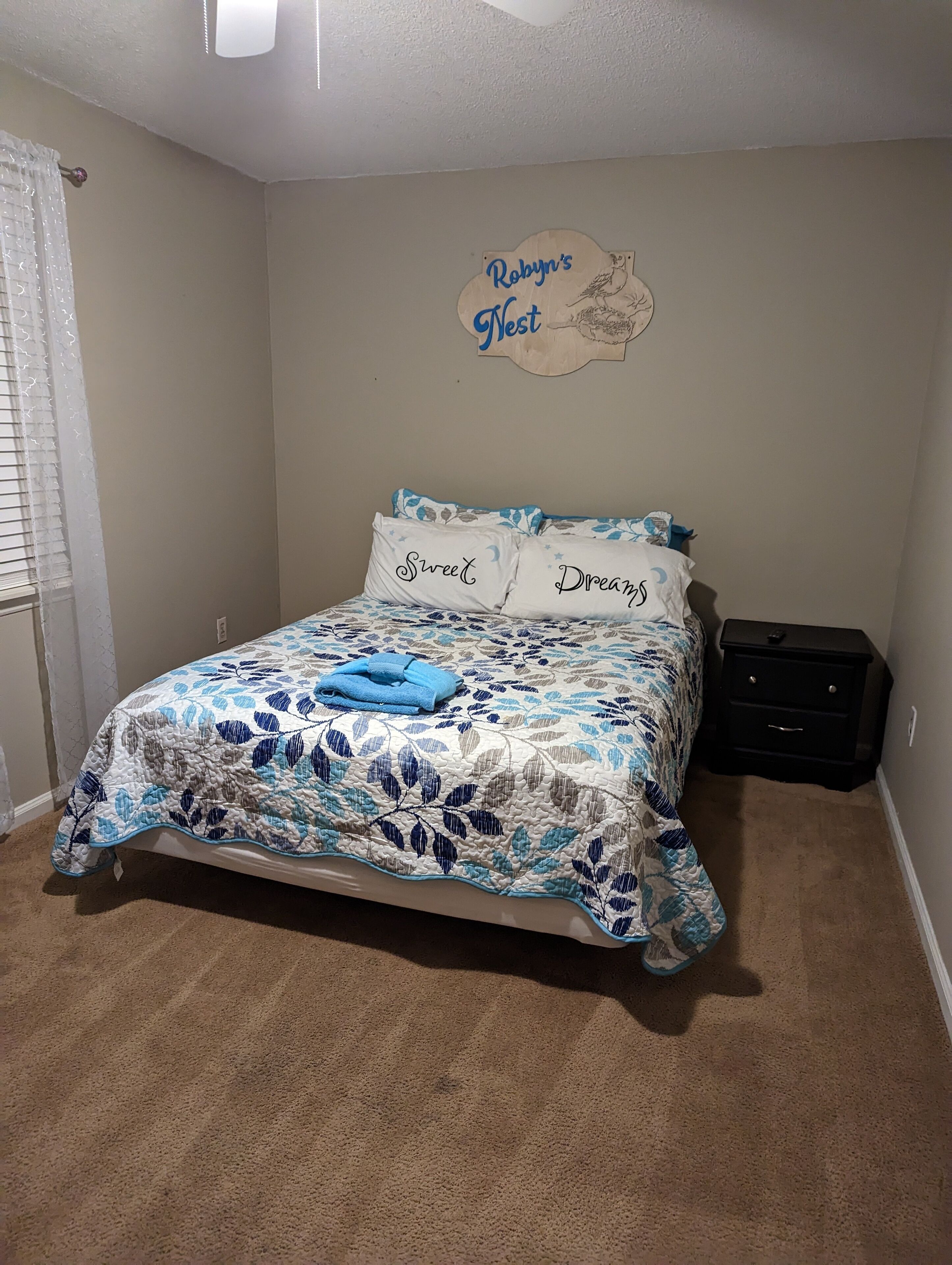 A bedroom with a bed, a wall sign, a nightstand, and a window with blinds.