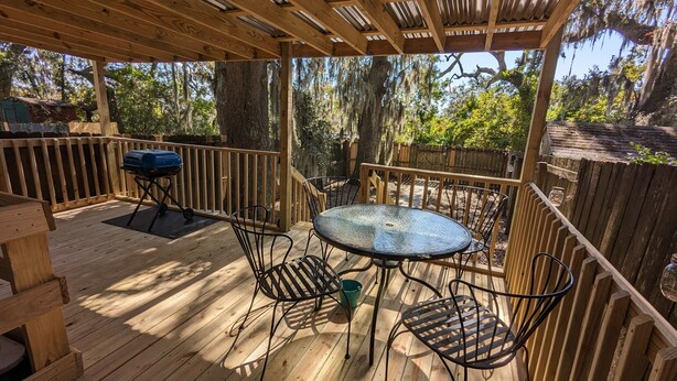 A wooden deck with a round glass table and metal chairs, surrounded by a wooden fence and trees. - Robyn's Nest (North Charleston)
