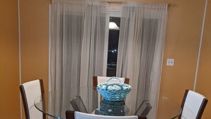 A dining area with a glass table surrounded by four chairs, a hanging light fixture, and a window with curtains. - Robyn's Nest (North Charleston)
