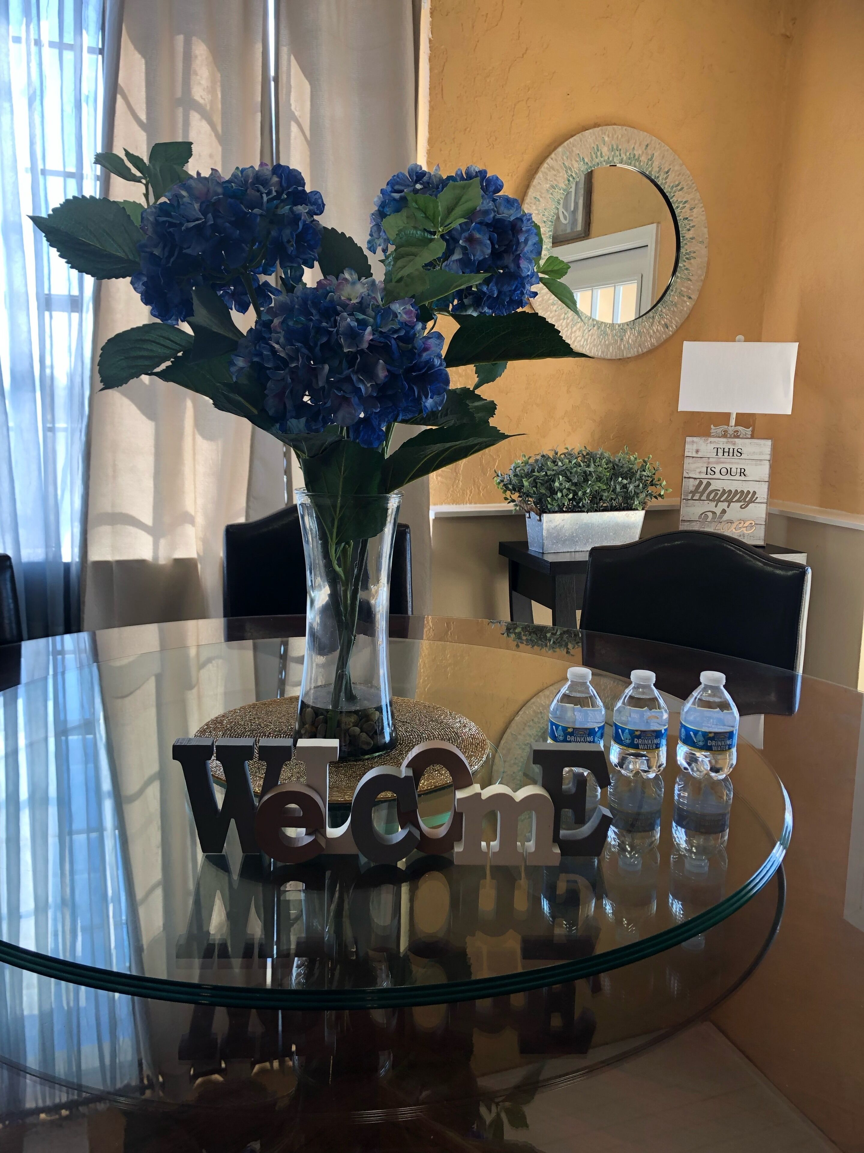 Une table à plateau en verre, un panneau d’accueil, un vase avec des fleurs bleues, une petite plante en pot et trois bouteilles d’eau.