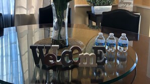 A glass-top table with a welcome sign, a vase with blue flowers, a small potted plant, and three water bottles.