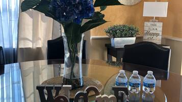 Une table à plateau en verre, un panneau d’accueil, un vase avec des fleurs bleues, une petite plante en pot et trois bouteilles d’eau.