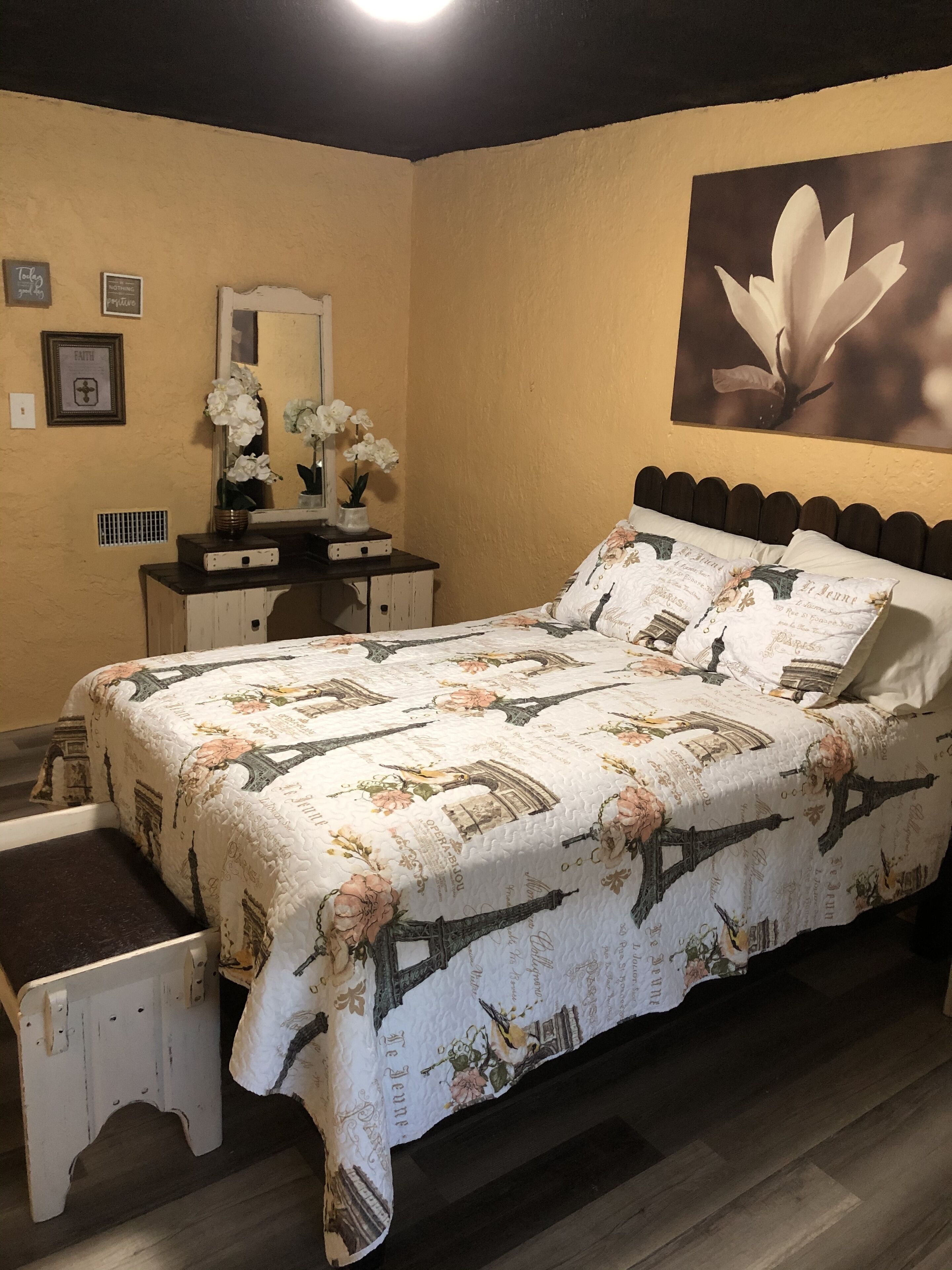 Un dormitorio con cama, cómoda, espejo y adornos de pared, incluyendo una pintura floral y objetos enmarcados.