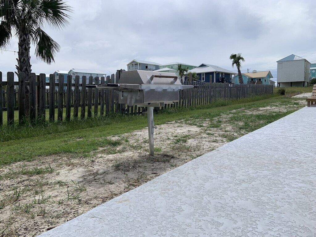 A beachside area with a barbecue grill, a wooden fence, and houses in the background.