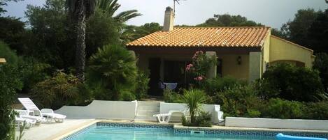 A pool area with lounge chairs, a building with a tiled roof, and surrounding greenery.
