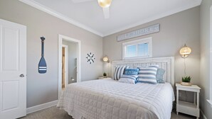 Une chambre avec un grand lit, un ventilateur de plafond, un panneau fixé au mur, une fenêtre avec des stores et une table de chevet avec une lampe.