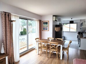 Un coin repas avec une table et des chaises en bois, une cuisine à l’arrière-plan et une vue sur l’extérieur grâce à de grandes fenêtres.