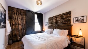 A bedroom with a wooden headboard, a large bed with white linens, a nightstand with a lamp, and a wall-mounted shelf. - The Rickhouse in NuLu~2 King Bed/2 Full Bath (Louisville)